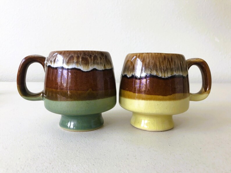 May include: Two ceramic mugs with brown, green, and yellow glaze. The mugs have a wide base and a rounded shape.
