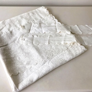 May include: White fabric with a detailed floral pattern and scalloped edges. The fabric is folded and appears to be a decorative element, possibly for a table setting or home decor. The overall aesthetic is delicate and elegant.
