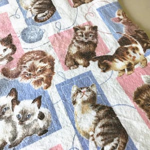 May include: A white fabric with a repeating pattern of various cat illustrations. The cats are depicted in different poses and colors, set against a backdrop of pink and blue squares. The overall theme is a playful and whimsical cat-themed design.