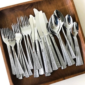 May include: A collection of stainless steel silverware, including forks, knives, and spoons, arranged in a wooden tray. The silverware has a textured handle design. The tray is square and made of dark wood.