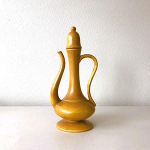 May include: A tall, mustard-yellow ceramic pitcher with a lid, curved spout, and handle. The pitcher has a bulbous base and a slender neck. The design is reminiscent of vintage or antique pottery.