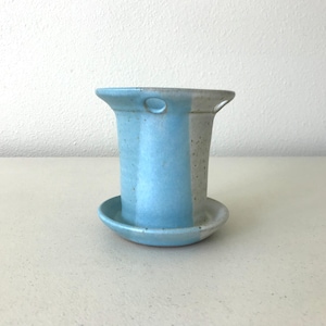 May include: A ceramic planter with a flared rim and an attached saucer base. The exterior features a vertical color-blocked design with alternating sections of light blue glaze and a speckled off-white finish, including two small circular holes.