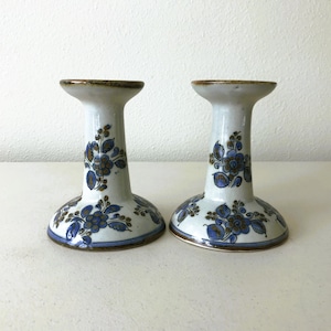 May include: Pair of ceramic candle holders. Each holder features a white base with blue floral designs and a brown rim. The candle holders are approximately 5 inches tall.