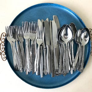 May include: A collection of assorted stainless steel silverware, including forks, knives, and spoons, arranged on a round, textured blue metal tray with decorative copper-colored handles on each side. The flatware features various patterns.