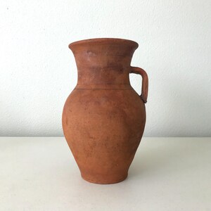 May include: A terracotta pitcher with a wide, flared rim and a single side handle. The vessel features a bulbous body that tapers toward the base, showcasing a natural, matte, reddish-brown clay texture with subtle variations in color throughout.