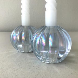 May include: Two round, ribbed glass candle holders with an iridescent finish, each holding a tall, white, spiral-textured taper candle. The glass bases feature a light blue tint that reflects rainbow-like colours across their curved, vertical surfaces.