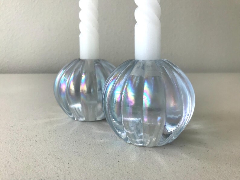 May include: Two round, ribbed glass candle holders with an iridescent finish, each holding a tall, white, spiral-textured taper candle. The glass bases feature a light blue tint that reflects rainbow-like colours across their curved, vertical surfaces.