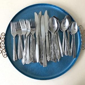 May include: A round blue metal serving tray with decorative gold-toned metal handles on each side. The tray contains a collection of assorted stainless steel flatware, including various forks, knives, and spoons with different handle patterns.