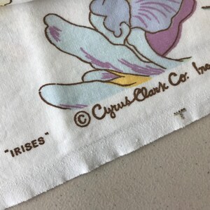 May include: Close-up of a white fabric with a floral iris pattern in shades of blue, purple, and yellow. The text "IRISES" and "Cyrus Clark Co. Inc." are printed on the fabric.