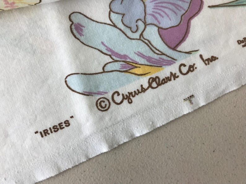 May include: Close-up of a white fabric with a floral iris pattern in shades of blue, purple, and yellow. The text "IRISES" and "Cyrus Clark Co. Inc." are printed on the fabric.