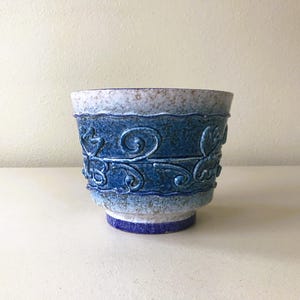 May include: A ceramic planter with a textured finish and a blue and white color scheme. The planter features a band of embossed floral scrollwork in a darker shade of blue. The base is a darker blue, and the rim is a lighter blue.