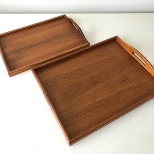 May include: Two rectangular wooden serving trays with a warm brown wood grain finish. Each tray features raised edges and integrated cutout handles on the shorter sides, showcasing a smooth surface and a consistent natural wood texture throughout.
