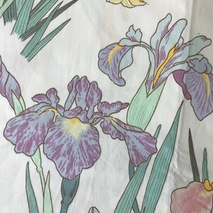 May include: Close-up of a fabric with a floral pattern featuring iris flowers. The irises are in shades of purple, lavender, and yellow, with green leaves and stems. The background is white, creating a contrast that highlights the flowers.