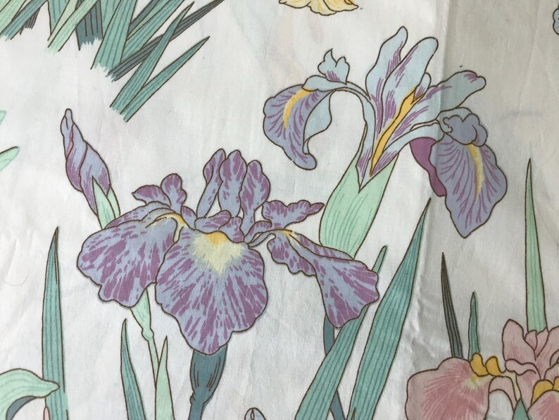May include: Close-up of a fabric with a floral pattern featuring iris flowers. The irises are in shades of purple, lavender, and yellow, with green leaves and stems. The background is white, creating a contrast that highlights the flowers.