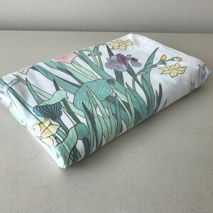 May include: Folded white fabric with a floral pattern featuring irises and other flowers in shades of pink, purple, and yellow, with green leaves and stems. The fabric is folded neatly, showcasing the design.
