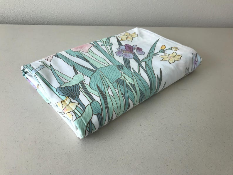 May include: Folded white fabric with a floral pattern featuring irises and other flowers in shades of pink, purple, and yellow, with green leaves and stems. The fabric is folded neatly, showcasing the design.