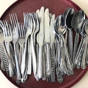 May include: A collection of assorted stainless steel flatware, including various forks, knives, and spoons with different handle patterns, resting on a round, dark red tray with decorative gold-toned metal handles on each side.