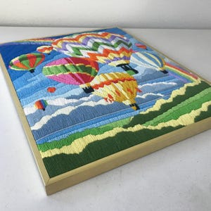 May include: Embroidered artwork featuring colorful hot air balloons floating in a blue sky with a rainbow. The framed piece has a gold-colored frame and depicts a landscape with green hills and white clouds. The artwork is a vibrant and cheerful piece.