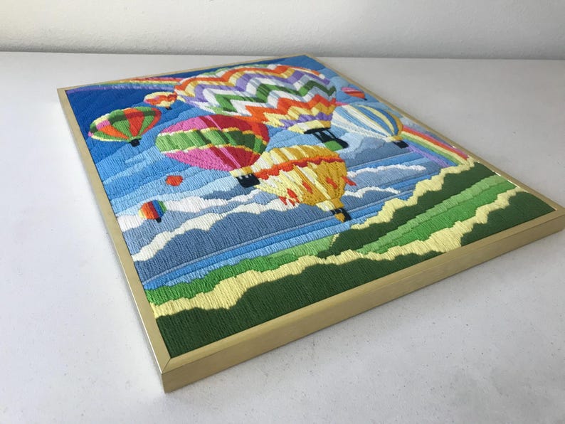 May include: Embroidered artwork featuring colorful hot air balloons floating in a blue sky with a rainbow. The framed piece has a gold-colored frame and depicts a landscape with green hills and white clouds. The artwork is a vibrant and cheerful piece.