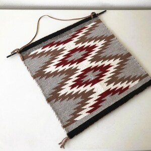 May include: A woven wall hanging with a geometric pattern in gray, white, brown, and burgundy. The rectangular textile is mounted on a black rod with a leather hanging strap. The design features zig-zag shapes.