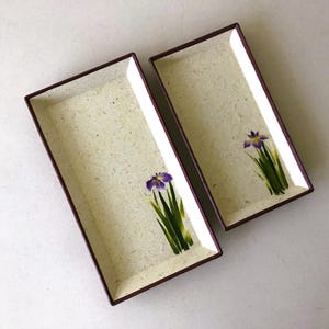 May include: Two rectangular serving trays with a light beige speckled surface and dark brown trim. Each tray features a hand-painted purple iris flower with green leaves. The trays are ideal for serving appetizers or sushi.