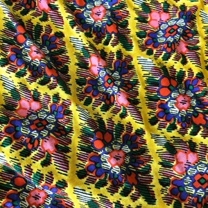 Op de afbeelding: Close-up van een levendige stof met een herhalend bloemenpatroon. Het ontwerp toont bloemen in roze, oranje en blauwe tinten tegen een heldergele achtergrond. De stof heeft een textuur met diagonale lijnen.