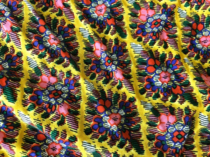 Op de afbeelding: Close-up van een levendige stof met een herhalend bloemenpatroon. Het ontwerp toont bloemen in roze, oranje en blauwe tinten tegen een heldergele achtergrond. De stof heeft een textuur met diagonale lijnen.