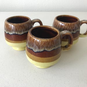 May include: Three brown and yellow ceramic mugs with a glazed finish. The mugs have a rounded shape and a handle on the side.