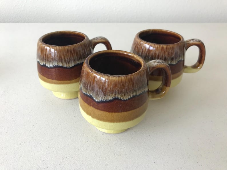 May include: Three brown and yellow ceramic mugs with a glazed finish. The mugs have a rounded shape and a handle on the side.