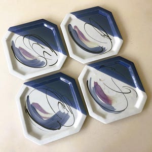 May include: Four octagonal ceramic plates with a white base, a blue border, and abstract purple and black brushstrokes. The plates are arranged in a square formation, showcasing a modern, artistic design.