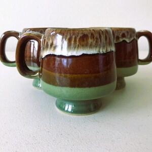May include: Set of three brown and green ceramic mugs with a white glaze. The mugs have a footed base and a rounded shape.