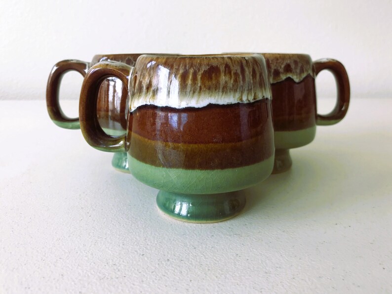 May include: Set of three brown and green ceramic mugs with a white glaze. The mugs have a footed base and a rounded shape.