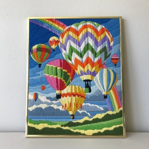 May include: Framed artwork featuring a colorful embroidered scene of hot air balloons floating in a blue sky with a rainbow. The balloons are various colors, including pink, green, yellow, and orange. The artwork is in a gold frame.