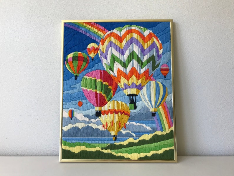 May include: Framed artwork featuring a colorful embroidered scene of hot air balloons floating in a blue sky with a rainbow. The balloons are various colors, including pink, green, yellow, and orange. The artwork is in a gold frame.