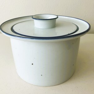 May include: A white ceramic lidded container with a speckled finish and a dark blue rim. The cylindrical vessel features a matching lid with a central knob handle, both accented by a thin dark blue line along the edges of the circular top.