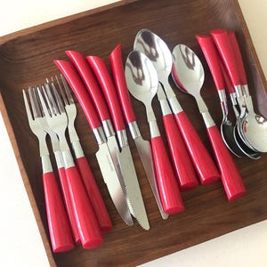 May include: A set of stainless steel flatware featuring bright red plastic handles. The collection includes three forks, three serrated knives, and four spoons, all arranged together. The utensils have a polished metallic finish and curved handles.