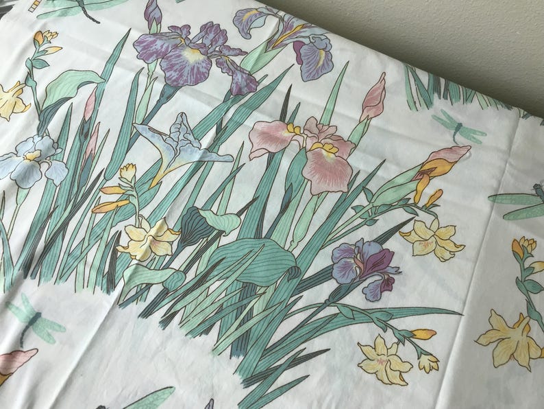 May include: White fabric with a floral pattern featuring irises in shades of purple, pink, and yellow, along with green leaves and stems. The design includes dragonflies. A soft, botanical theme.