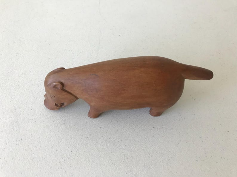 K&ouml;nnte beinhalten: Holzskulptur eines Capybaras mit polierter Oberfl&auml;che. Die braune Figur hat einen abgerundeten K&ouml;rper, kurze Beine und einen kleinen Schwanz. Das Capybara ist in einer entspannten Pose dargestellt, mit leicht gesenktem Kopf.