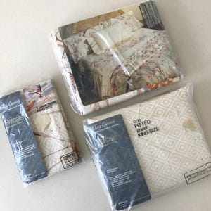 May include: Three packages of Oleg Cassini king size bedding in plastic wrap. One package shows a floral bed set, one contains two pillowcases with a geometric pattern, and one contains a fitted sheet with a white and beige diamond grid design.