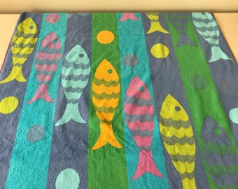 May include: A rectangular beach towel featuring a repeating pattern of stylized fish in yellow, pink, and light blue, set against vertical stripes of blue, teal, and green. The design includes scattered solid circles in matching colors.