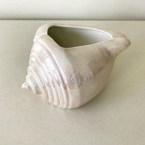 May include: A white ceramic seashell planter with a pearlescent finish. The planter is shaped like a seashell with a wide opening at the top.