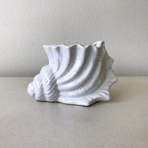 May include: A white ceramic seashell-shaped decorative planter. The planter has a textured surface with detailed ridges and a wide opening. The seashell is resting on a light-colored surface, suitable for coastal or nautical decor.