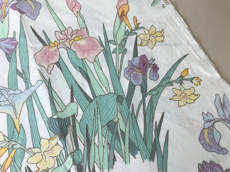 May include: White fabric with a floral pattern featuring irises in shades of pink, purple, and yellow, with green leaves and stems. The design has a vintage, hand-drawn aesthetic, suitable for fabric or home decor.