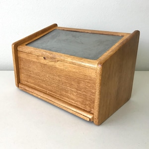 May include: A rectangular wooden bread box with a light brown natural wood grain finish. The box features a slanted top lid with a rectangular gray panel insert and a small horizontal cutout handle on the front panel for opening the lid.