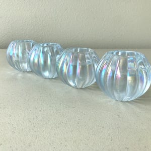 May include: A set of four small, round glass bowls featuring a vertical ribbed texture and an iridescent finish that reflects light with subtle rainbow hues. Each bowl has a wide, open top and a curved, bulbous shape, creating a uniform appearance.