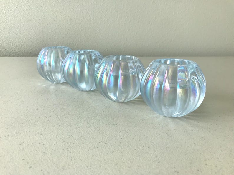 May include: A set of four small, round glass bowls featuring a vertical ribbed texture and an iridescent finish that reflects light with subtle rainbow hues. Each bowl has a wide, open top and a curved, bulbous shape, creating a uniform appearance.