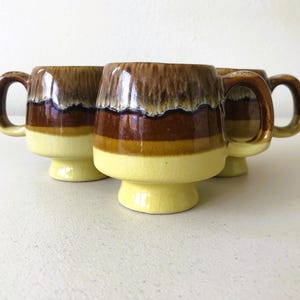 May include: Set of four ceramic mugs with a brown, yellow, and white glaze. The mugs have a wide base and a rounded shape.