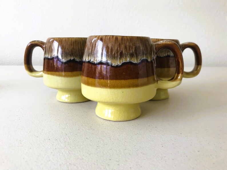 May include: Set of four ceramic mugs with a brown, yellow, and white glaze. The mugs have a wide base and a rounded shape.