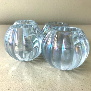 May include: A set of three small, spherical glass candle holders featuring a vertical ribbed texture and an iridescent finish that reflects light with subtle blue and purple hues. Each piece has a wide circular opening at the top.