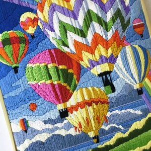 May include: A framed embroidery featuring colorful hot air balloons against a blue sky with clouds. The balloons are various colors, including pink, green, yellow, and orange. The artwork has a detailed, textured appearance.
